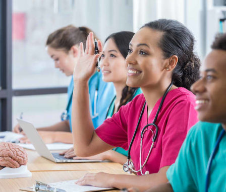 Smiling young female African American student raises her hand to ask a question in class. She is sitting in the middle of three other students at a string of desks. Two other students are looking forward and smiling and another is writing at her desk.